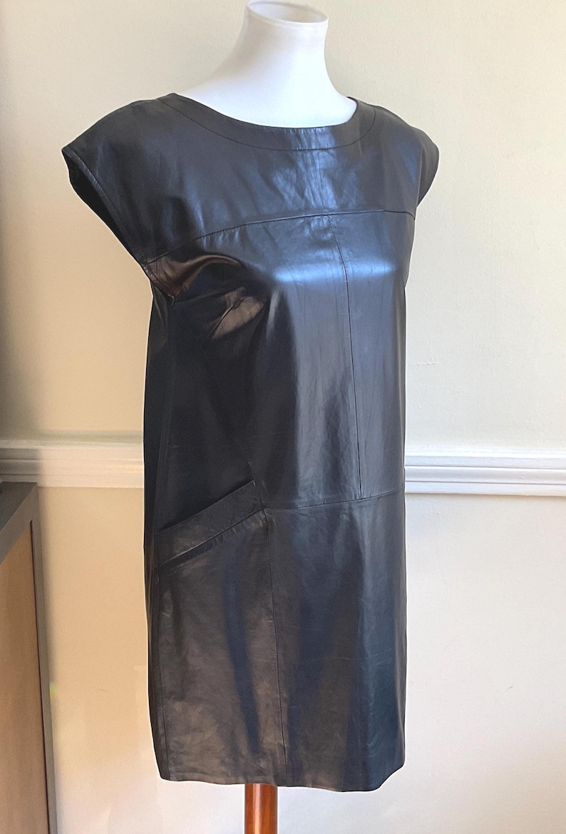 May include: A black leather dress with a round neckline and short sleeves. The dress has a straight silhouette and a single pocket on the front. The dress is displayed on a mannequin.