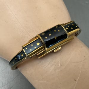 May include: A gold-tone bangle watch featuring a black enamel inlay decorated with small gold stars and dots. The watch has a rectangular case with a side-mounted winding crown and a hinged design that allows the band to open and close securely.