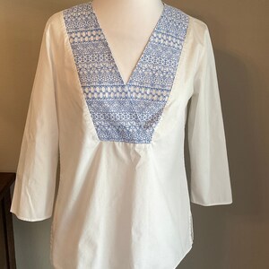 May include: White cotton blouse with a blue embroidered neckline. The embroidery features a geometric pattern.