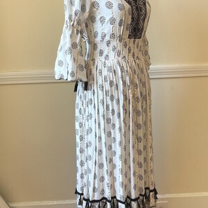 May include: White maxi dress with a black floral pattern and a V-neckline. The dress features a smocked waist, three-quarter sleeves, and black tassel trim. The dress is displayed on a black mannequin.