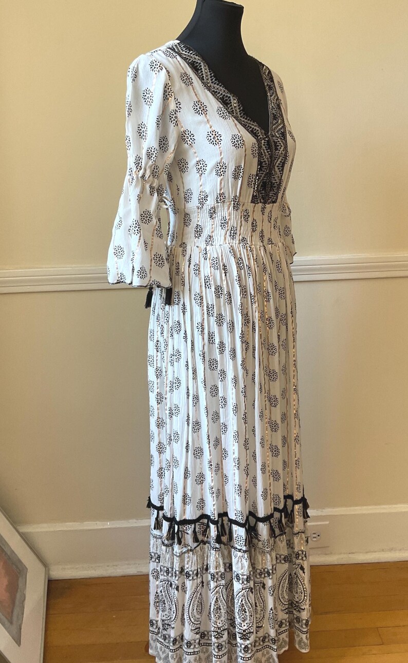 May include: White maxi dress with a black floral pattern and a V-neckline. The dress features a smocked waist, three-quarter sleeves, and black tassel trim. The dress is displayed on a black mannequin.