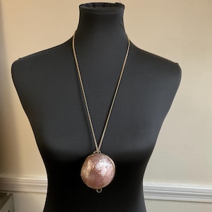 May include: A gold-toned chain necklace with a large, round pendant. The pendant is a light pink color with a textured, cracked surface. The necklace is displayed on a black mannequin.