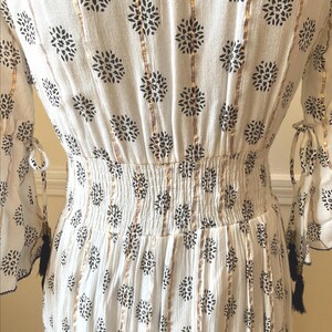 May include: White dress with black floral pattern and gold vertical stripes. The dress features a shirred waist, long sleeves with tie details, and black tassels. The fabric has a crinkled texture.