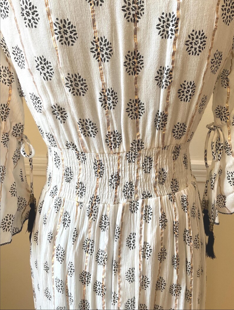 May include: White dress with black floral pattern and gold vertical stripes. The dress features a shirred waist, long sleeves with tie details, and black tassels. The fabric has a crinkled texture.