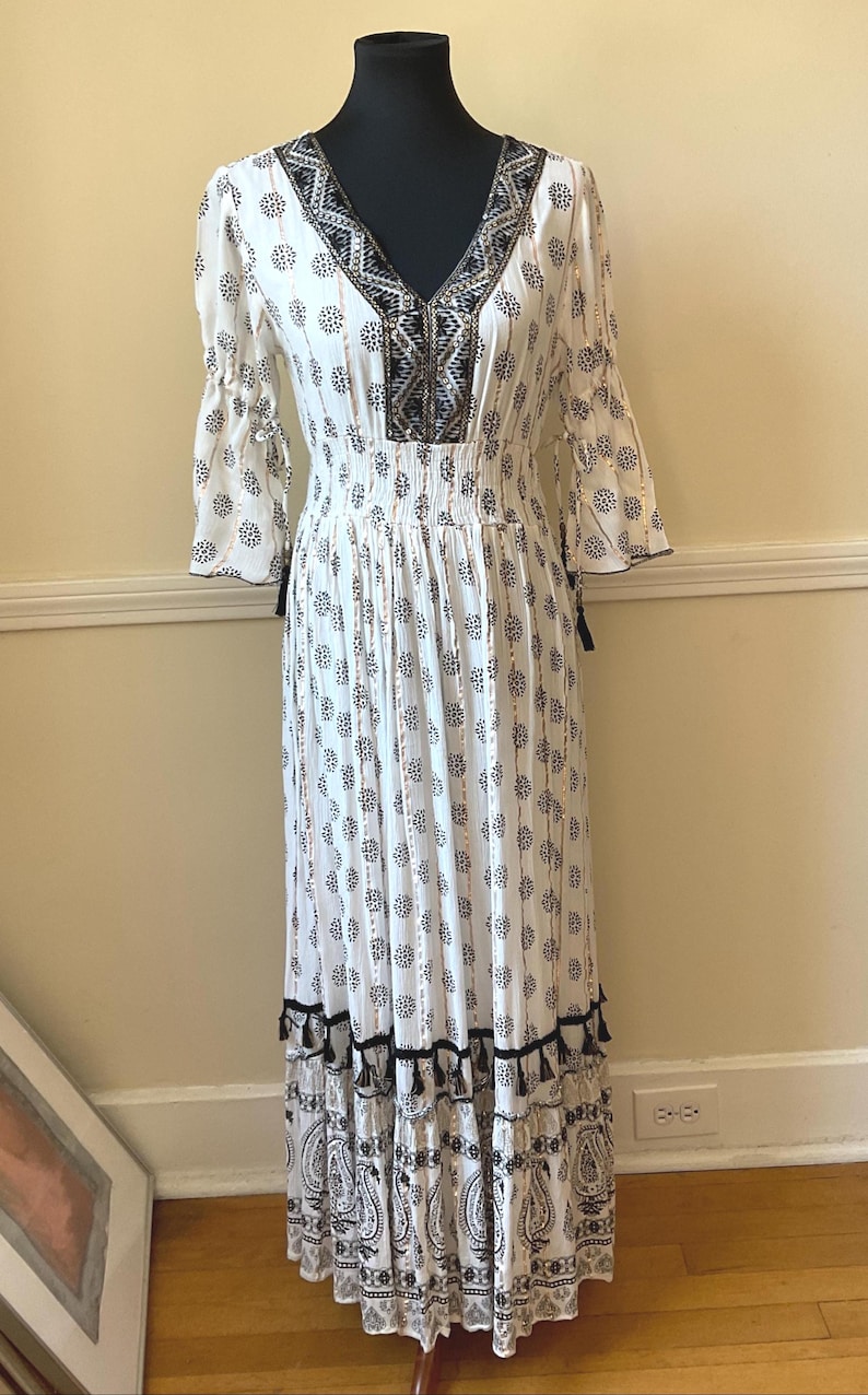 May include: White maxi dress with a V-neck and three-quarter sleeves. The dress features a black and white floral pattern, a smocked waist, and black tassel accents. The hemline has a paisley print.
