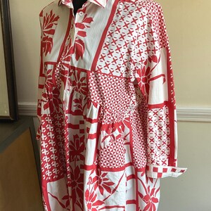 May include: A long-sleeved shirt dress featuring a white base with a bold red patchwork-style print. The pattern includes various geometric shapes, floral motifs, and cross-hatch designs. It has a classic collar, a button-down front, and cuffs.