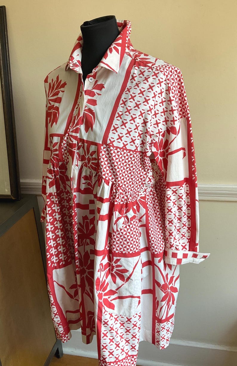 May include: A long-sleeved shirt dress featuring a white base with a bold red patchwork-style print. The pattern includes various geometric shapes, floral motifs, and cross-hatch designs. It has a classic collar, a button-down front, and cuffs.