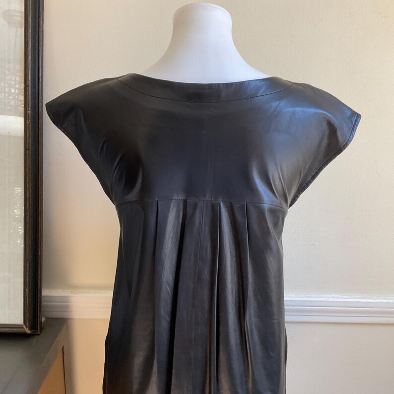 May include: A black leather dress with a scoop neckline and cap sleeves. The dress has vertical pleats below the bust and is displayed on a mannequin. The dress has a sleek, minimalist design.