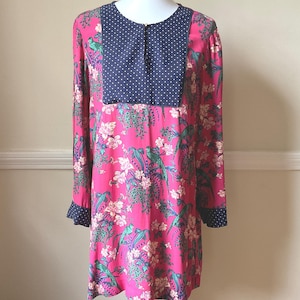 May include: A long-sleeved tunic dress in a vibrant pink hue, adorned with a floral and bird pattern. The dress features a navy blue polka-dot yoke and cuffs. The tunic is displayed on a mannequin.