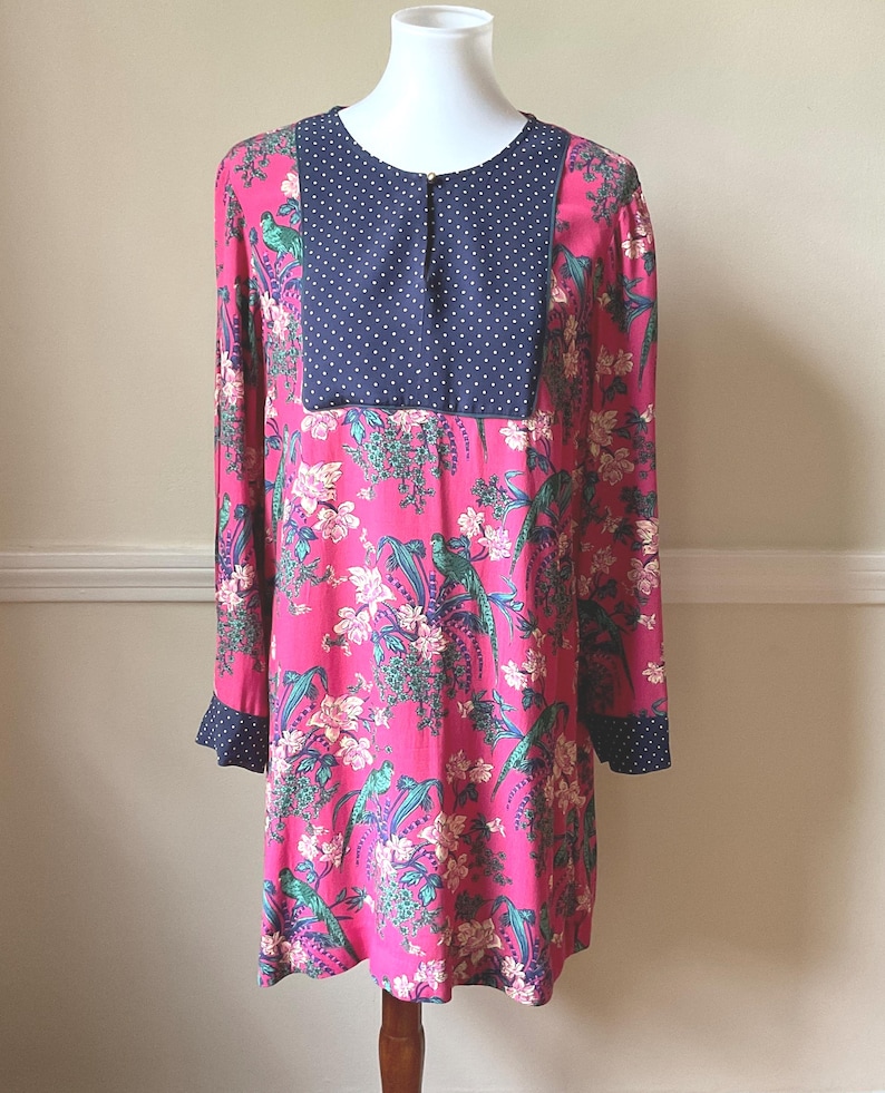 May include: A long-sleeved tunic dress in a vibrant pink hue, adorned with a floral and bird pattern. The dress features a navy blue polka-dot yoke and cuffs. The tunic is displayed on a mannequin.