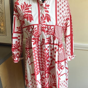 May include: A long-sleeved shirt dress featuring a white base with a bold red patchwork-style print. The design includes various geometric patterns, floral motifs, and leaf shapes. It has a collared neckline, a button-down front, and a gathered waist.