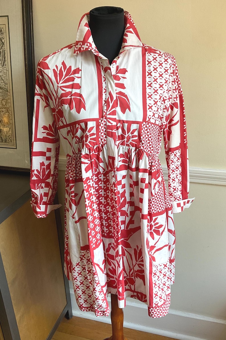 May include: A long-sleeved shirt dress featuring a white base with a bold red patchwork-style print. The design includes various geometric patterns, floral motifs, and leaf shapes. It has a collared neckline, a button-down front, and a gathered waist.