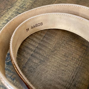 Puede incluir: Un cintur&oacute;n de cuero beige enrollado con costuras visibles a lo largo de los bordes. El interior del cintur&oacute;n presenta el texto estampado M 44828 en una fuente oscura, indicando un identificador de modelo o talla en la superficie lisa.