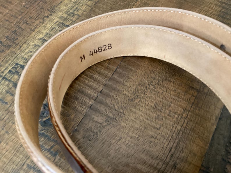 Puede incluir: Un cintur&oacute;n de cuero beige enrollado con costuras visibles a lo largo de los bordes. El interior del cintur&oacute;n presenta el texto estampado M 44828 en una fuente oscura, indicando un identificador de modelo o talla en la superficie lisa.