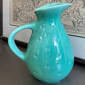 May include: A turquoise ceramic pitcher with a glossy finish and a textured surface featuring raised circular dots. The pitcher has a curved handle on the side and a wide, open spout at the top, showcasing a rounded body and a smooth, uniform color.