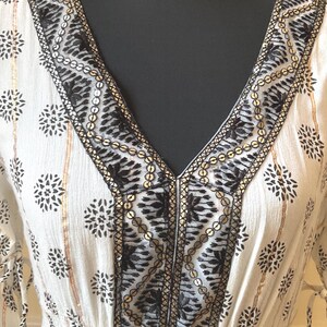 May include: White V-neck dress with black floral patterns and gold accents. The neckline and sleeves are adorned with intricate black embroidery and gold sequins. The dress has a fitted waist and long sleeves.