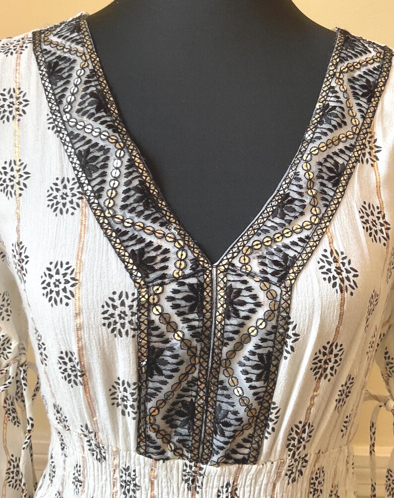 May include: White V-neck dress with black floral patterns and gold accents. The neckline and sleeves are adorned with intricate black embroidery and gold sequins. The dress has a fitted waist and long sleeves.