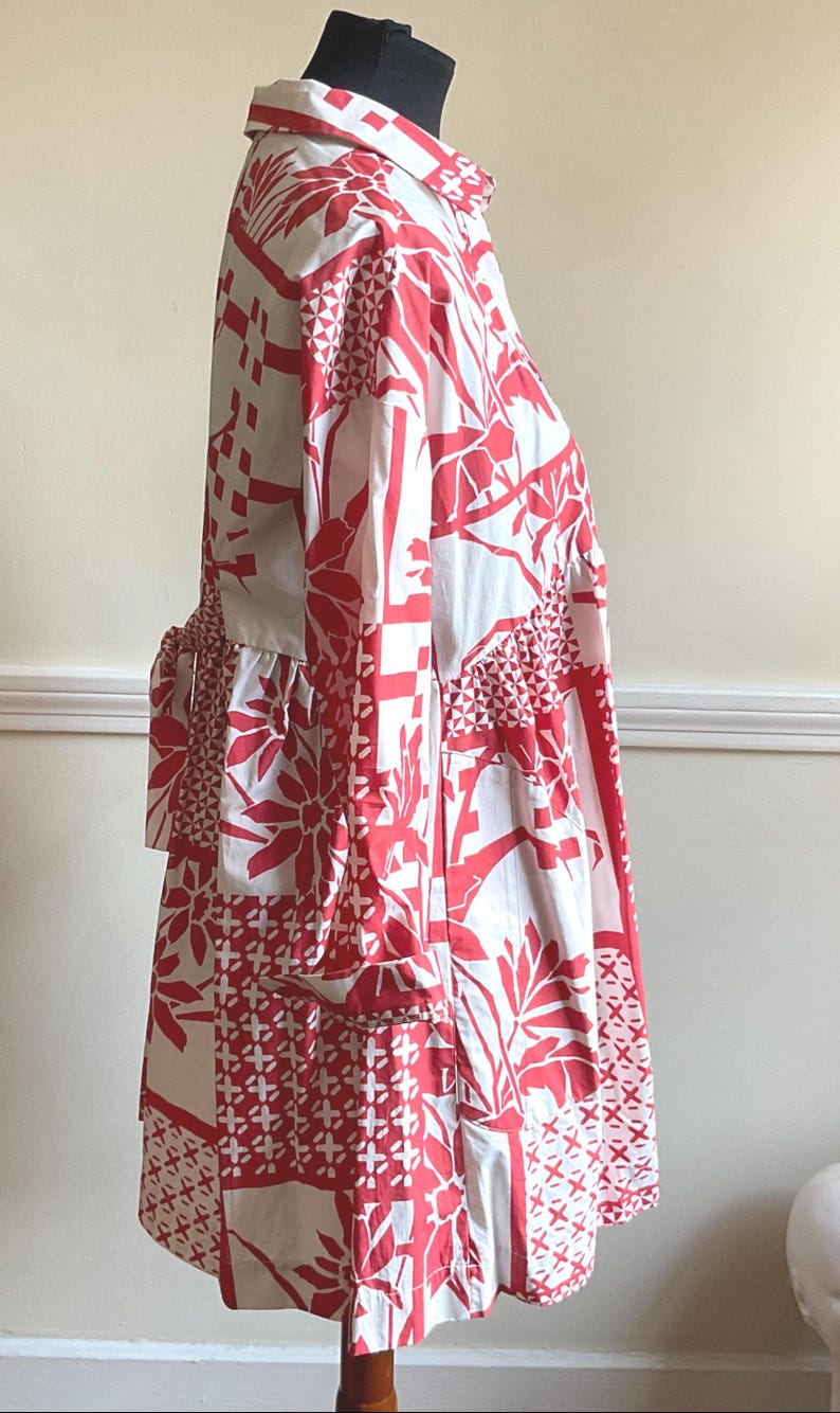 May include: A long-sleeved shirt dress featuring a white base with a bold red graphic pattern of geometric shapes, floral motifs, and cross-hatch designs. The garment includes a classic collar, a button-down front, and a tie-waist detail at the back.