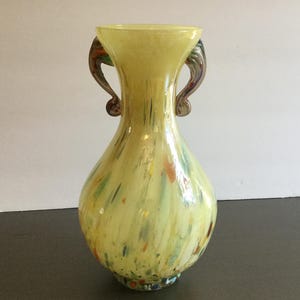 May include: A yellow glass vase with two curved handles. The vase has a speckled design with colorful accents of blue, orange, and green. The vase is a decorative item.