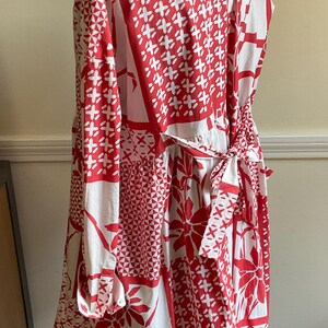 May include: A long-sleeved shirt dress featuring a white and red patchwork-style print with floral and geometric patterns. The garment includes a collared neckline, buttoned cuffs, and a fabric tie belt at the waist that is tied in a bow at the back.