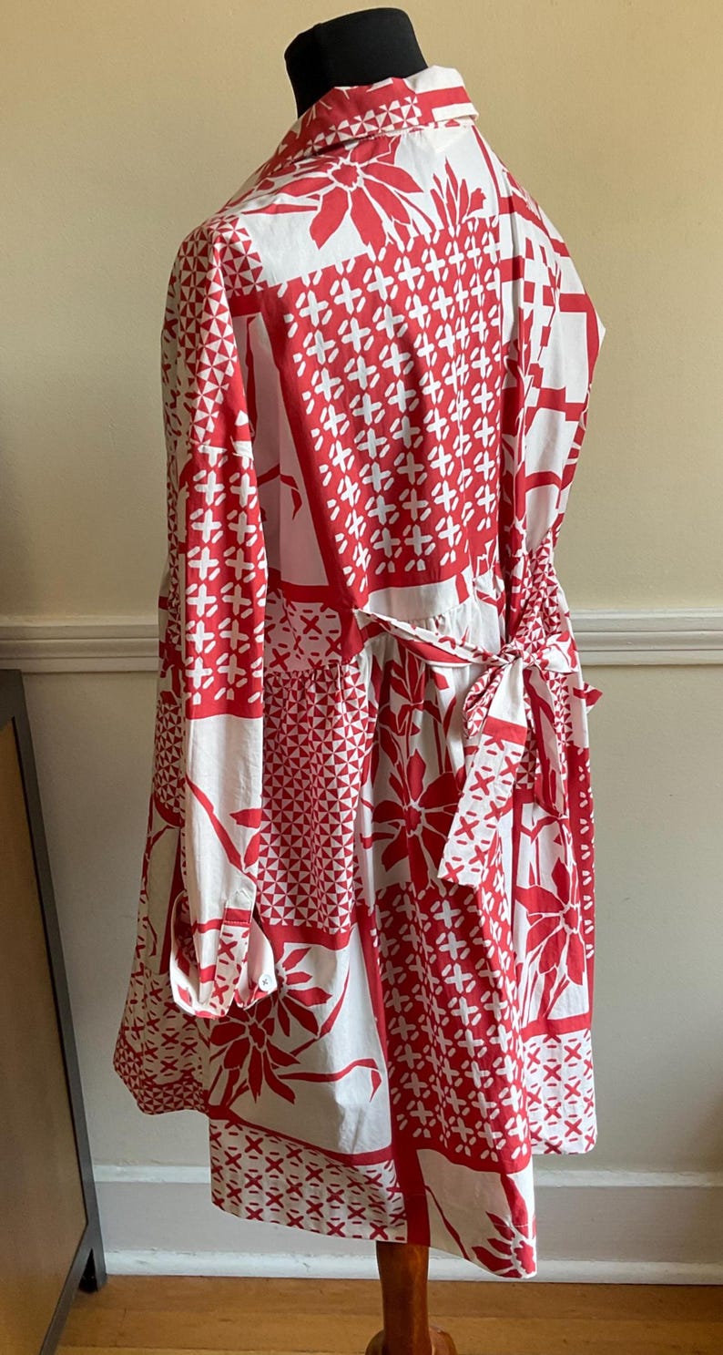May include: A long-sleeved shirt dress featuring a white and red patchwork-style print with floral and geometric patterns. The garment includes a collared neckline, buttoned cuffs, and a fabric tie belt at the waist that is tied in a bow at the back.