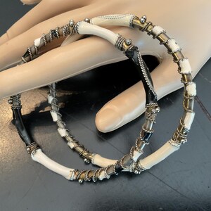 May include: Two white and black bangle bracelets with gold and silver accents. The bracelets are made of bone and have a woven design.