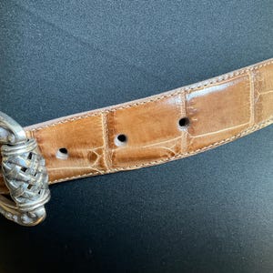 Puede incluir: Un cintur&oacute;n de color tostado con una textura brillante de piel de cocodrilo en relieve y una hebilla de metal plateado pulido. La hebilla presenta un dise&ntilde;o central trenzado complejo con detalles ornamentales de metal en el marco.