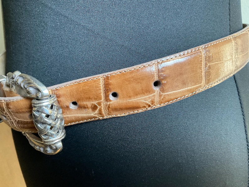 Puede incluir: Un cintur&oacute;n de color tostado con una textura brillante de piel de cocodrilo en relieve y una hebilla de metal plateado pulido. La hebilla presenta un dise&ntilde;o central trenzado complejo con detalles ornamentales de metal en el marco.