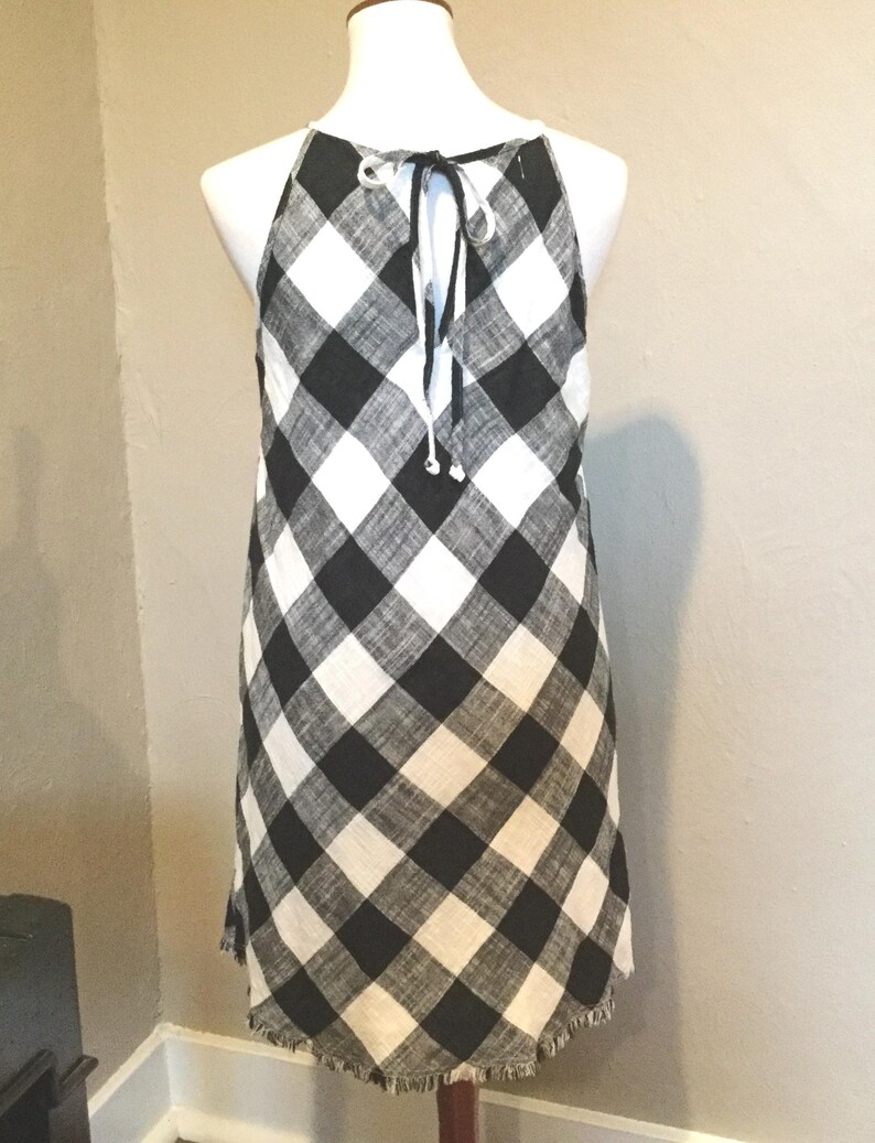 May include: A black and white checkered halter dress with a frayed hem. The dress is displayed on a mannequin. The dress has a tie closure at the back of the neck.