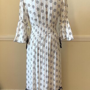 May include: White maxi dress with a floral pattern in black and gold. The dress features a V-neck, elastic waist, and bell sleeves. Black tassels adorn the sleeves and hemline, adding a bohemian flair to the garment.