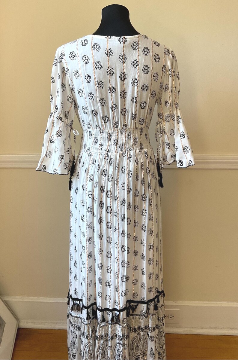 May include: White maxi dress with a floral pattern in black and gold. The dress features a V-neck, elastic waist, and bell sleeves. Black tassels adorn the sleeves and hemline, adding a bohemian flair to the garment.