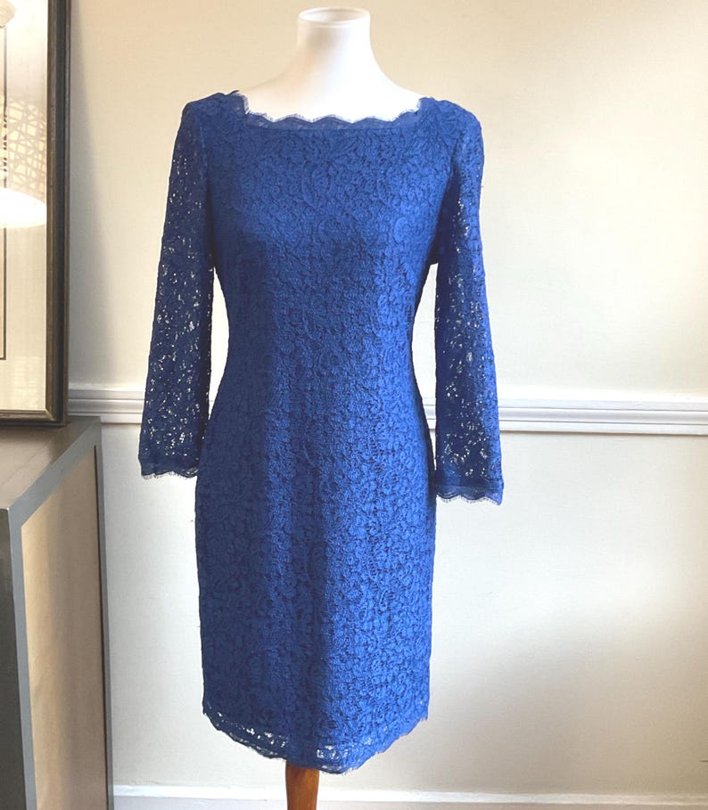 May include: A cobalt blue lace dress with long sleeves and a scalloped neckline. The dress is displayed on a mannequin. The dress is knee-length and has a fitted silhouette.