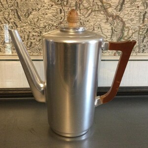 May include: A tall, cylindrical silver-toned metal coffee pot featuring a long, angled spout, a flat lid with a small, rounded wooden knob handle, and a dark brown wooden handle attached to the side with metallic hardware.