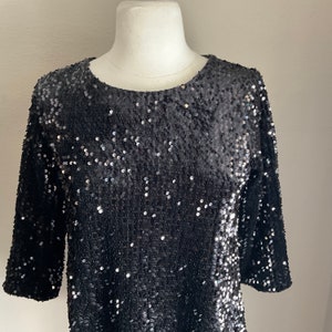 May include: A black sequin dress with a round neckline and three-quarter sleeves. The dress is displayed on a mannequin. The sequins create a shimmering effect, reflecting light. The dress is a formal or party wear item.