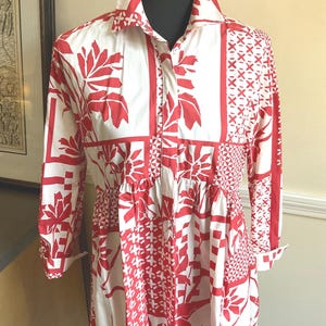 May include: A long-sleeved shirt dress featuring a white base with a bold red graphic pattern. The design includes panels of floral motifs, geometric shapes, and cross-hatch details. It has a classic collar, a button-down front, and a gathered waist.