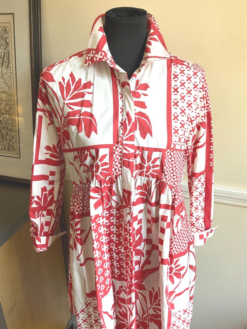 May include: A long-sleeved shirt dress featuring a white base with a bold red graphic pattern. The design includes panels of floral motifs, geometric shapes, and cross-hatch details. It has a classic collar, a button-down front, and a gathered waist.