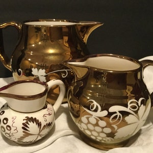 May include: Three gold-colored ceramic pitchers of varying sizes. The pitchers have a glossy finish and are decorated with white grape and leaf designs. The smallest pitcher has a white body with a maroon rim and handle.