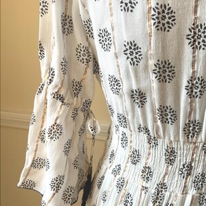 May include: White dress with black floral pattern and gold vertical stripes. The dress features flared sleeves with tie details and a gathered waist. The fabric has a textured appearance.