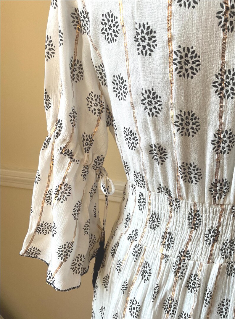 May include: White dress with black floral pattern and gold vertical stripes. The dress features flared sleeves with tie details and a gathered waist. The fabric has a textured appearance.