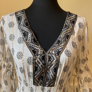 May include: A white dress with a V-neckline, adorned with black floral patterns and gold accents. The dress features a cinched waist and flowing sleeves, creating an elegant and bohemian look. The neckline is embellished with black and gold sequins.