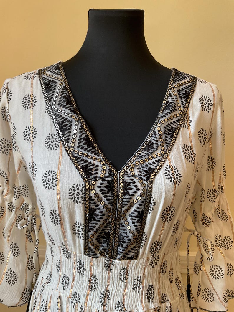 May include: A white dress with a V-neckline, adorned with black floral patterns and gold accents. The dress features a cinched waist and flowing sleeves, creating an elegant and bohemian look. The neckline is embellished with black and gold sequins.