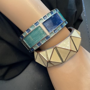 May include: Two decorative bangles. The top bangle features rectangular sections of blue and teal enamel with silver-colored metal borders. The bottom bangle has a geometric design with cream-colored enamel and copper-colored metal accents.