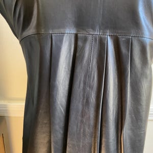 May include: Close-up of a dark grey leather dress. The dress has a pleated design on the front, with vertical seams and a subtle sheen. The garment appears sleeveless, with a simple, elegant silhouette.