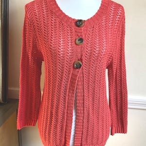 May include: Coral-colored open-front cardigan with three large brown buttons. The lightweight knit sweater has a round neckline and three-quarter sleeves. The open weave design adds a delicate texture to the garment. The cardigan is displayed on a mannequin.