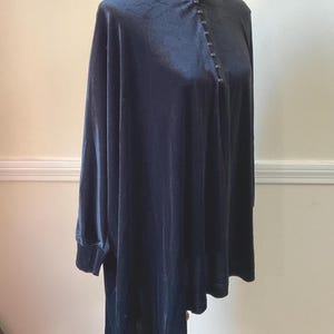May include: A dark navy blue velvet tunic with a high, button-up collar and long sleeves. The tunic has a slightly asymmetrical hemline and is displayed on a wooden dress form. The fabric has a soft sheen.