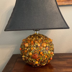 May include: A table lamp with a black lampshade and a round, mosaic base made of various shades of brown, orange, and gold. The base is covered in small pieces of tile, shells, and other decorative elements.