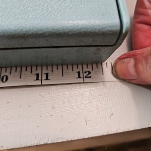 Peut inclure: Une boîte bleu clair texturée mesurant 30,48 cm. La boîte est mesurée avec un ruban à mesurer blanc. La boîte a une forme rectangulaire et semble être faite d'un matériau dur.