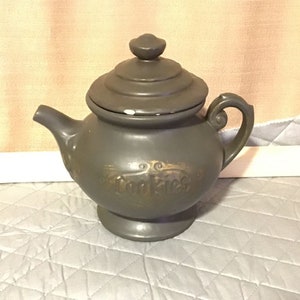 Puede incluir: Una galleta de cerámica gris en forma de tetera, con tapa y asa. La palabra "Cookies" está escrita en letras doradas en la parte delantera. La jarra tiene un pico y se asienta sobre una pequeña base.