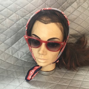 May include: A mannequin head wearing red cat-eye sunglasses and a patterned headscarf. The sunglasses have a bold, retro style. The headscarf features a red, white, and blue design.