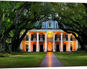 May include: A white antebellum mansion with a large porch and columns, surrounded by a canopy of lush green trees. The house is lit up at night, creating a warm and inviting atmosphere.